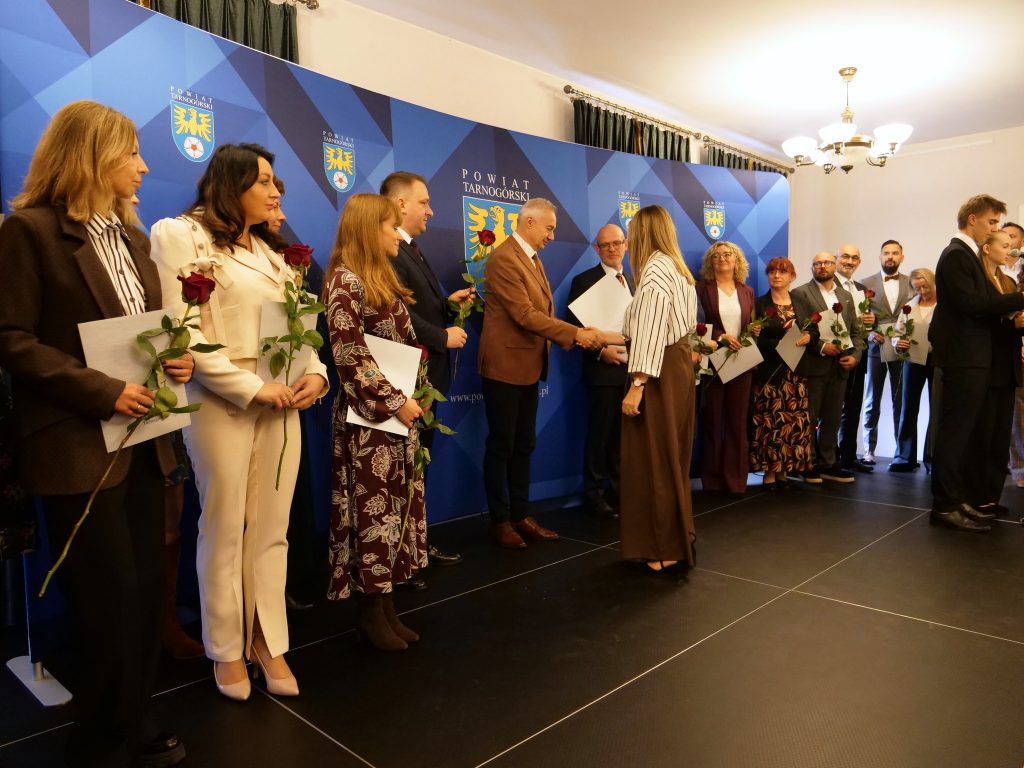 grupa osób na scenie, mężczyzna uściska kobiecie dłoń, reszta osób stoi na scenie z różami i dyplomami w rękach