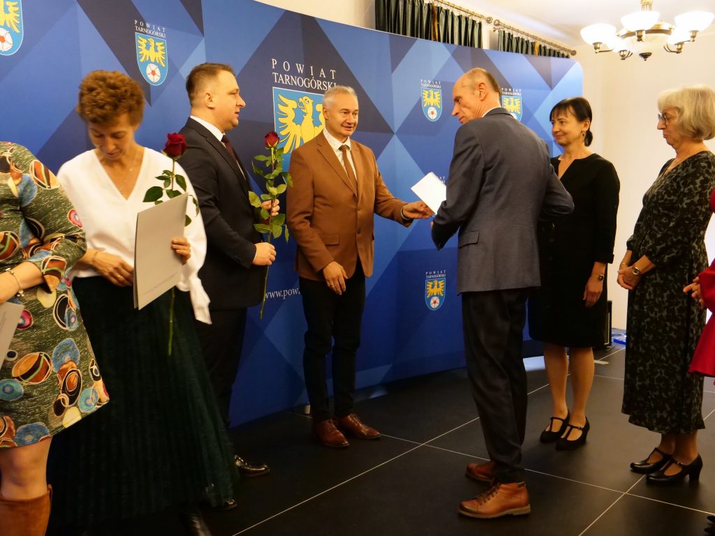 mężczyzna wręcza innemu mężczyźnie dokument, obok mężczyzna trzyma w ręku różę, obok kobieta trzyma w ręku dyplom i różę, w oddali stoją dwie kobiety