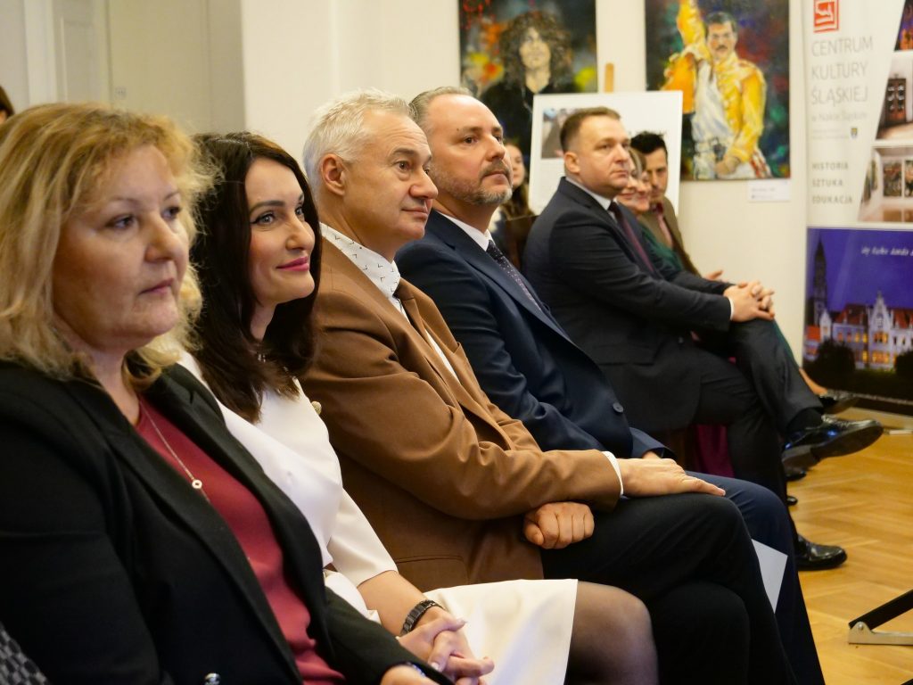 kobiety i mężczyźni elegancko ubrani siedzą w rzędzie na widowni, zwróceni w prawą stronę, w tle na ścianie wiszą obrazy