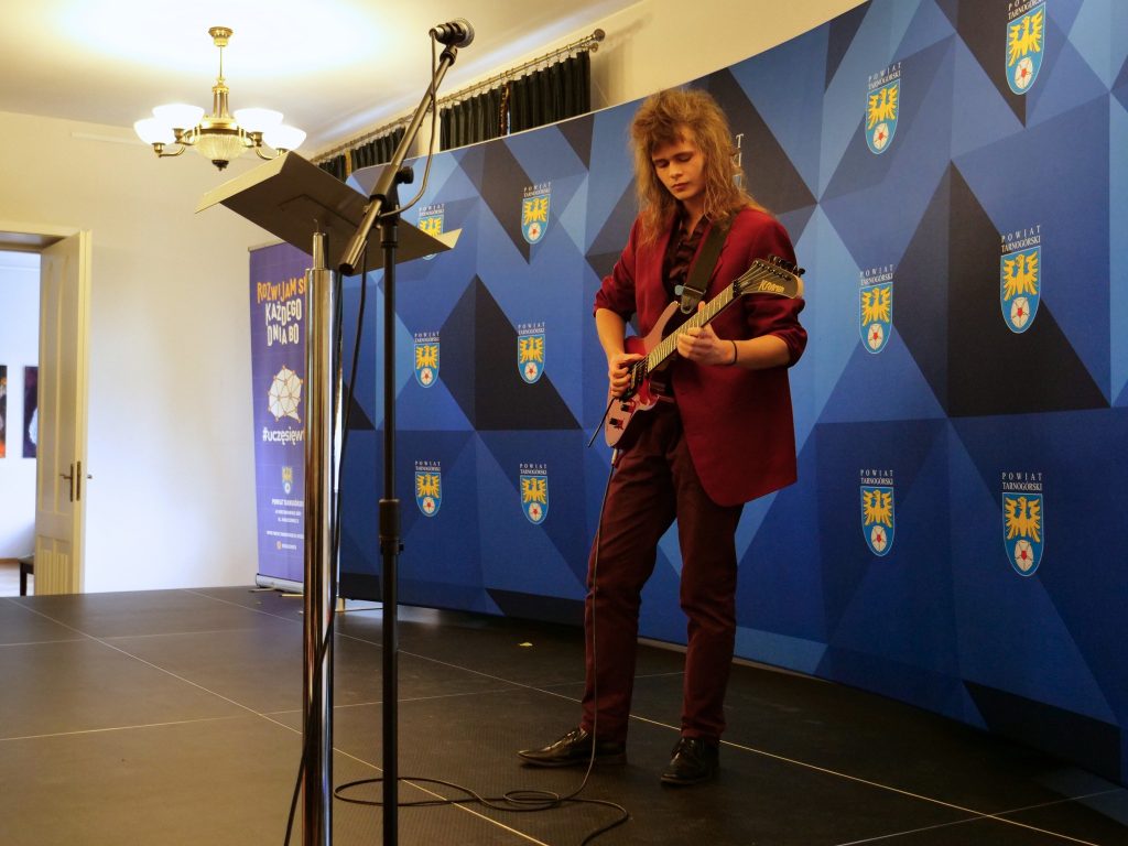 na scenie chłopak grający na gitarze elektrycznej, w tle ścianka z herbami powiatu tarnogórskiego, przed nim mikrofon