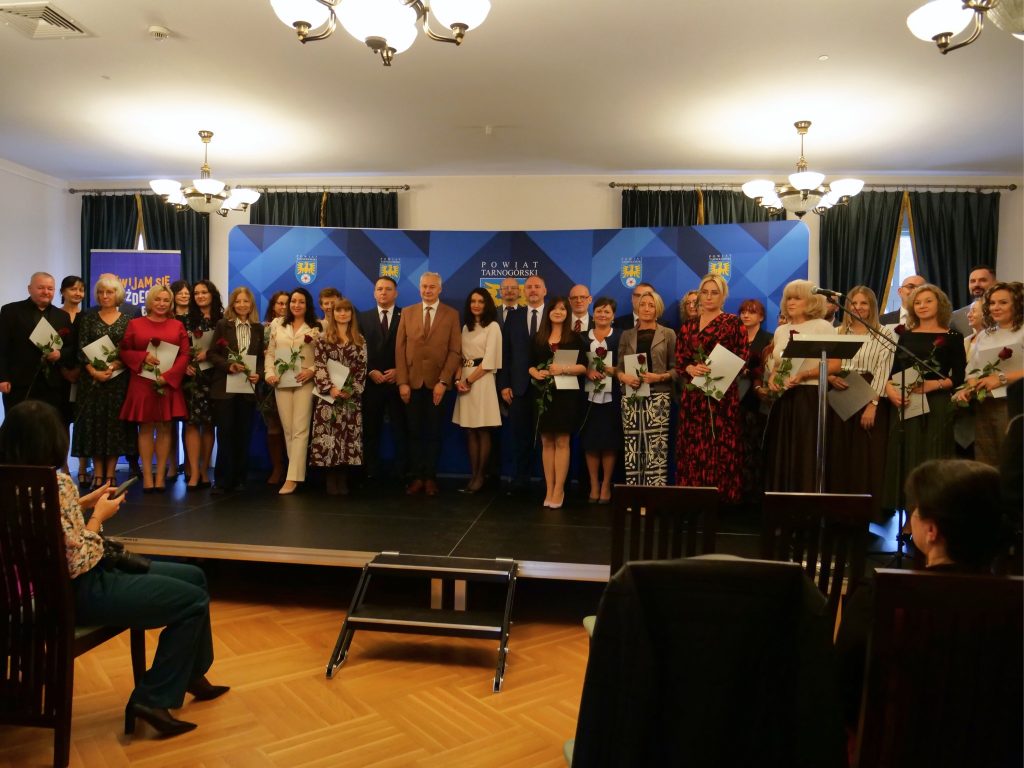 grupa osób stoi na scenie, pozuje do zdjęcia, w rękach trzymają różę i dyplomy, z przodu widoczna widownia i osoby na niej siedzące