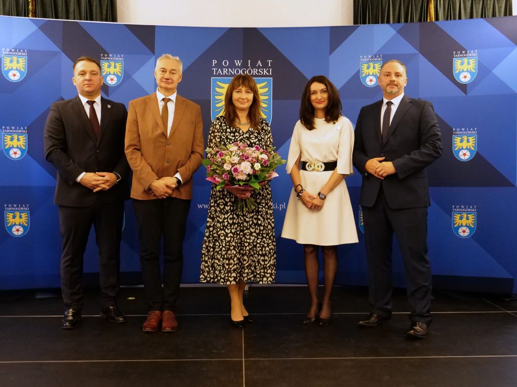 trzech mężczyzn i dwie kobiety pozują do zdjęcia na ściance z herbami powiatu tarnogórskiego, jedna z kobiet w ręku trzyma bukiet kwiatów