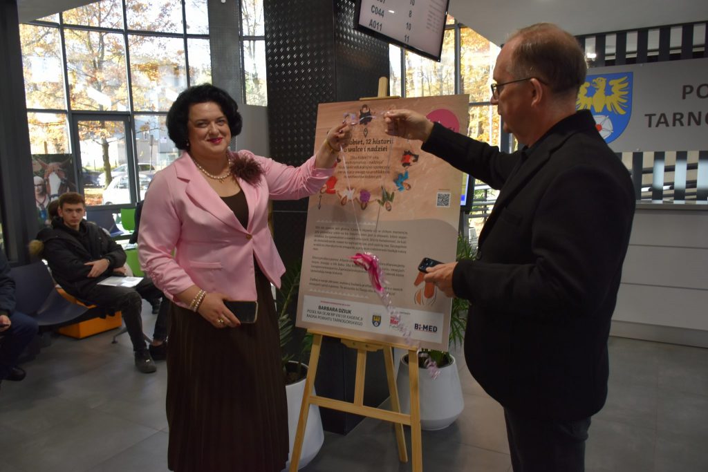 kobita elegancko ubrana i mężczyzna w garniturze, między nimi obraz z napisami, w rękach trzymają wstążkę