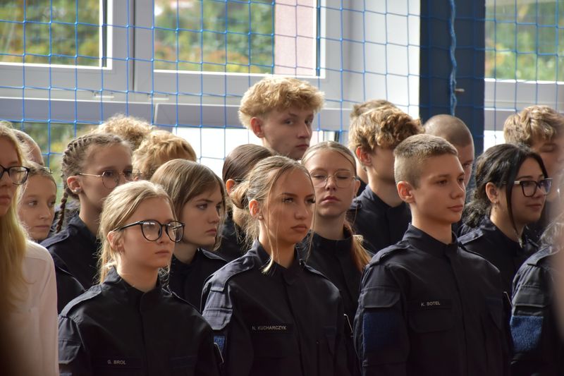 grupa uczniów w mundurach z klasy policyjnej, za nimi siatka i okna