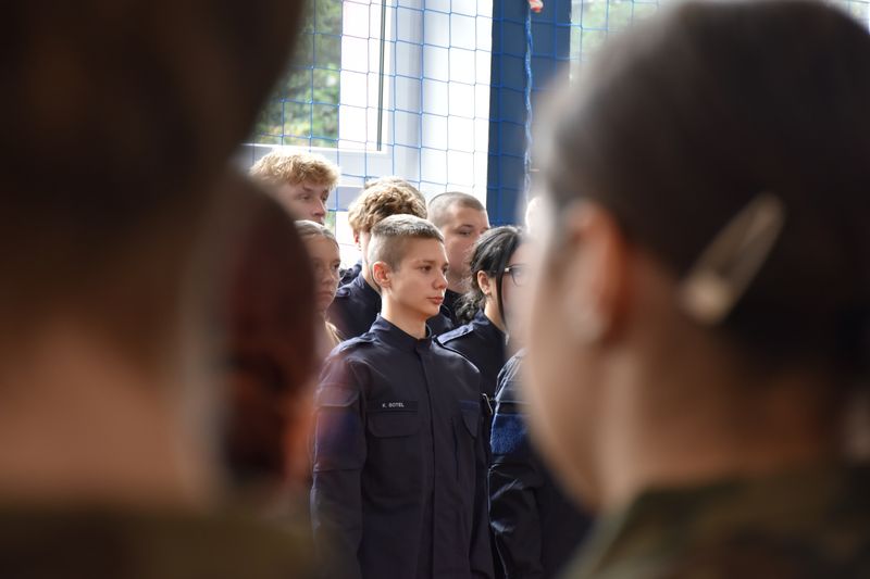 na pierwszym planie dwie zamazane głowy uczniów, na drugim planie uczniowie stoją w szeregu w mundurach