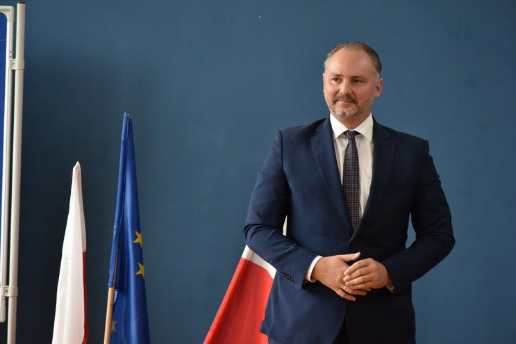 mężczyzna w garniturze, stoi i ma założone ręce, obok flagi polski i flaga europejska