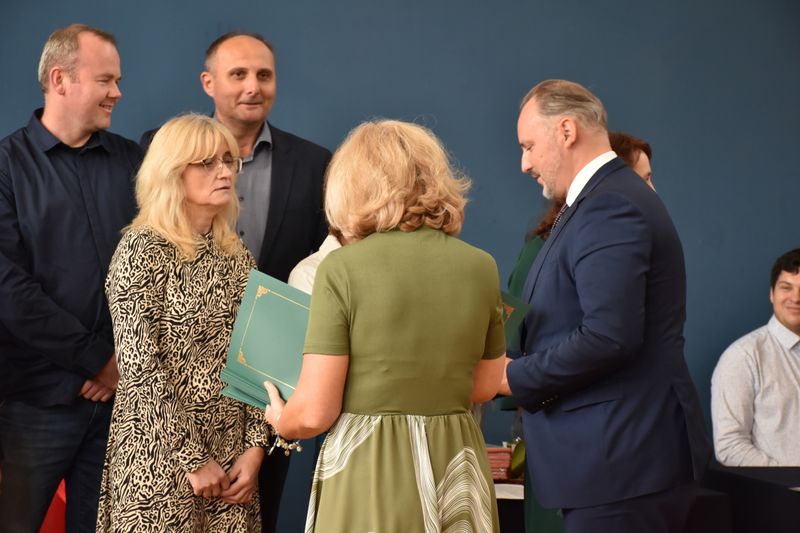 kobieta trzyma teczki, mężczyzna obok wręcza je drugiej kobiecie, obok dwóch mężczyzn elegancko ubranych