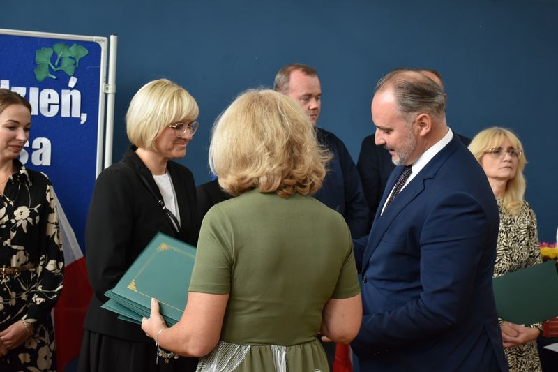 Kobieta elegancko ubrana w rękach trzyma uroczyste teczki, obok mężczyzna w garniturze, obok stoją inne osoby uroczyście ubrane.