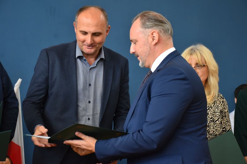 mężczyzna w garniturze wręcza dyplom drugiemu mężczyźnie w garniturze, obok fragment kobiety