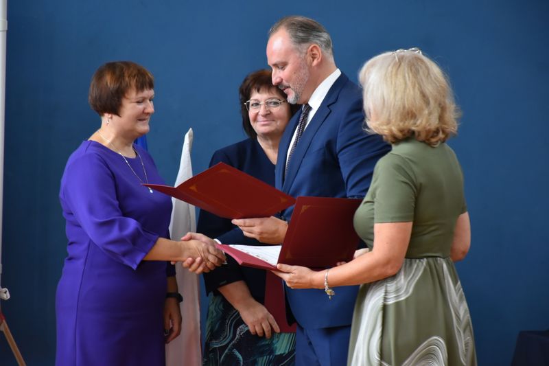 mężczyzna w garniturze wręcza kobiecie dokument, druga kobieta stoi obok niego trzymając w rękach dokument, w tle fragment kobiety