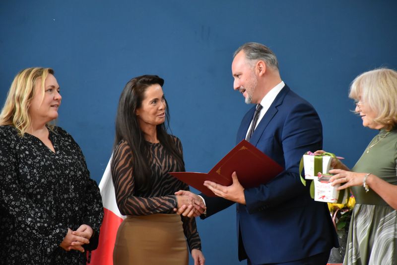 mężczyzna w garniturze wręcza kobiecie teczkę z dokumentem, miedzy nimi dowie kobiety, kobita po prawej trzyma w ręku upominek
