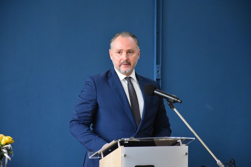 mężczyzna w garniturze stoi za mównicą, przed nim mikrofon, w tle ściana i fragment bukietu kwiatów
