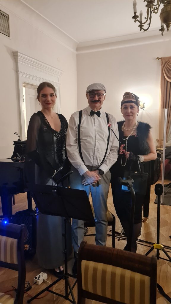 dwie kobiety ubrane na styl lat 20, w środku przebrany mężczyzna, w tle pianino, instrumenty 