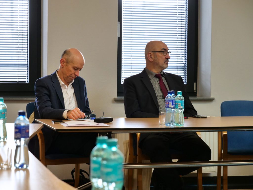 dwóch mężczyzn, jeden z nich ma przed sobą dokumenty, spogląda w dół, drugi patrzy w lewo