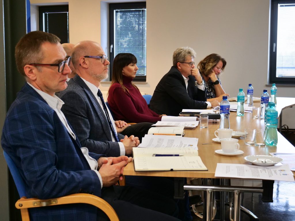 mężczyźni elegancko ubrani i kobiety elegancko ubrane siedzą przy stole, na stole dokumenty, butelki z wodą i filiżanki, w tle okna