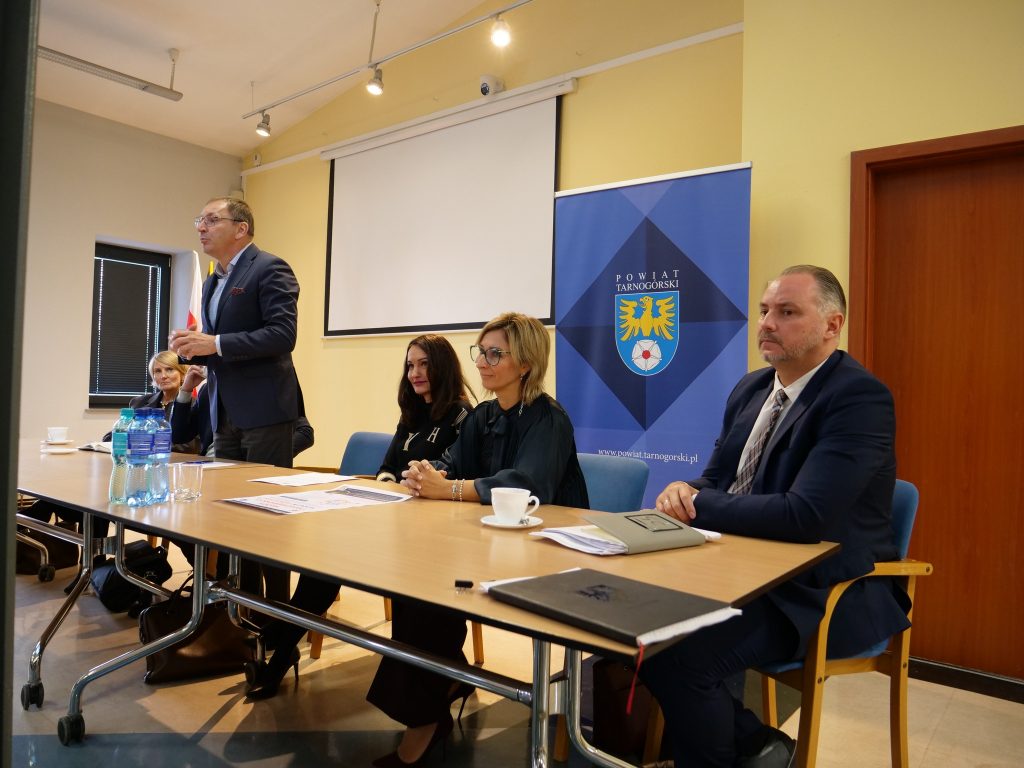 pięć osób siedzi przy stole, jedna osoba stoi przy stole i ma złożone ręce, na stole butelki z wodą i dokumenty