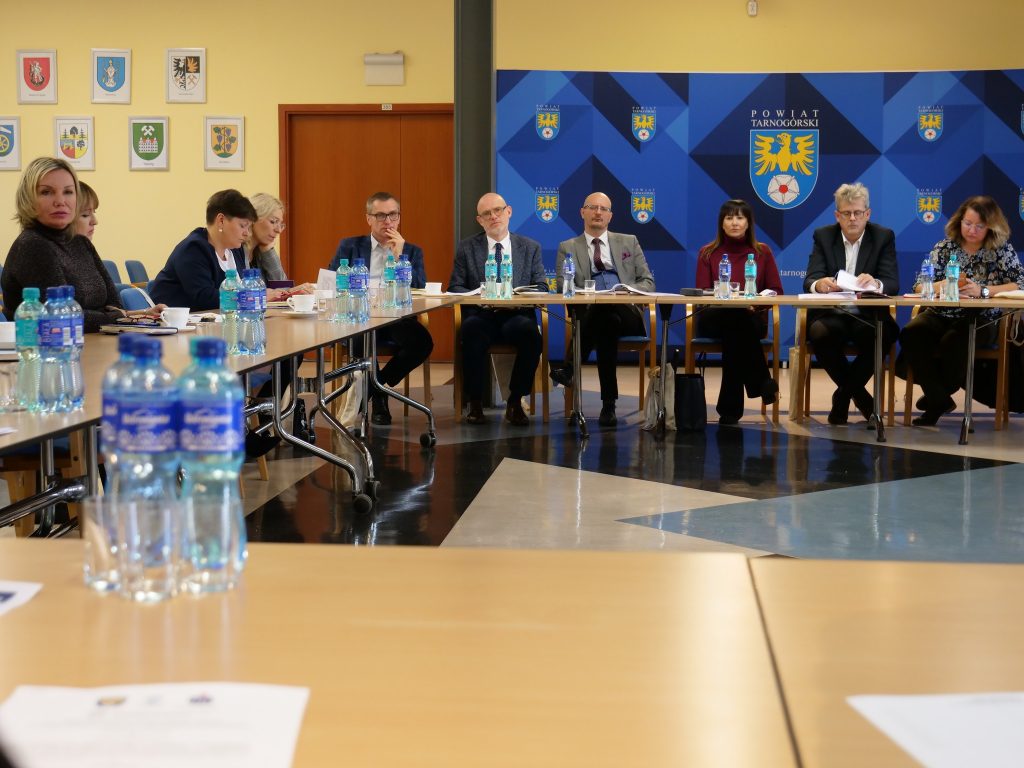 grupa osób, siedzą przy stole, na stole butelki z wodą, filiżanki i dokumenty, w tle ścianka z herbem powiatu tarnogórskiego