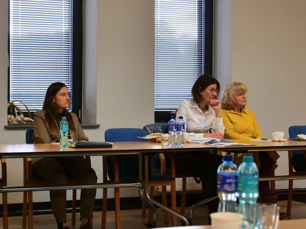 kobiety, siedzą przy stole, zwrócone w lewą stronę, na stole butelki z wodą, w tle okna z żaluzjami