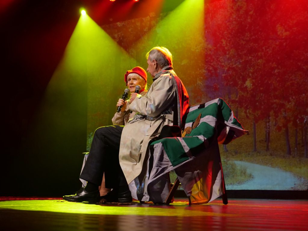 dwie osoby, na scenie, przebrane, siedzą na kanapie, w tle kolorowe lampy 