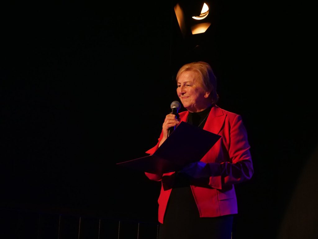 uśmiechnięta kobieta na scenie, w rękach trzyma mikrofon i teczkę