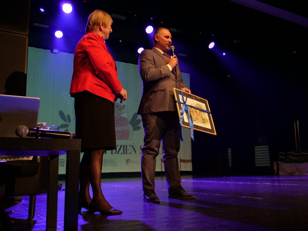 kobieta na scenie, obok mężczyzna trzyma w ręce mikron a w drugiej obraz, po lewej stronie fragment fortepianu 