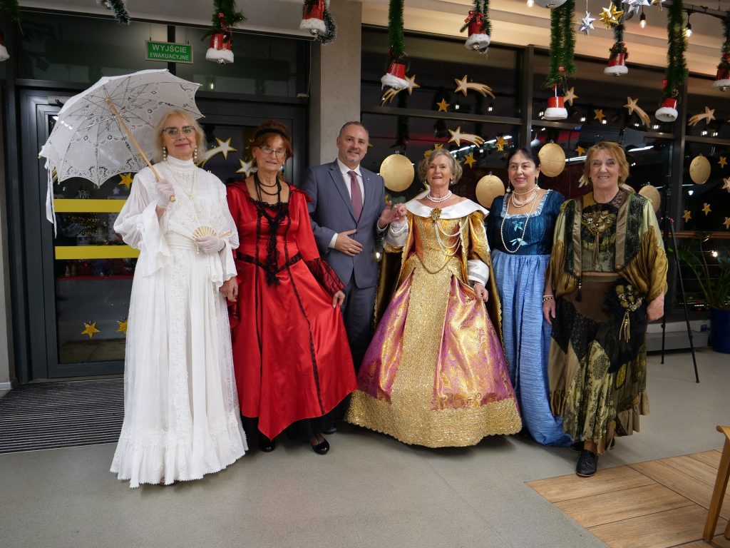 pięć kobiet ubrane w stroje historyczne, między nimi mężczyzna w garniturze, w tle okna i drzwi automatyczne 
