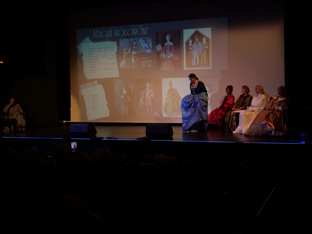 na scenie kobiety w strojach historycznych, w tle ekran multimedialny na którym wyświetlana jest prezentacja obrazków