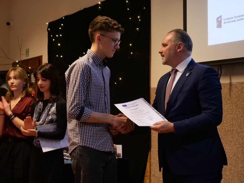 mężczyzna w garniturze wręczna jednemu z uczniów dyplom, obok stoją uczennice i klaszczą w dłonie