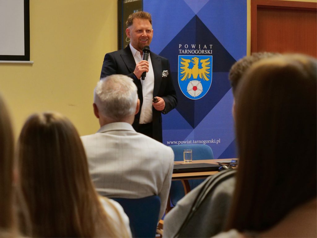 mężczyzna, ubrany w garnitur, trzyma w ręku mikrofon, w tle kolorowa ścianka i fragment ekranu multimedialnego, z przodu fragment osób, siedzących na widowni