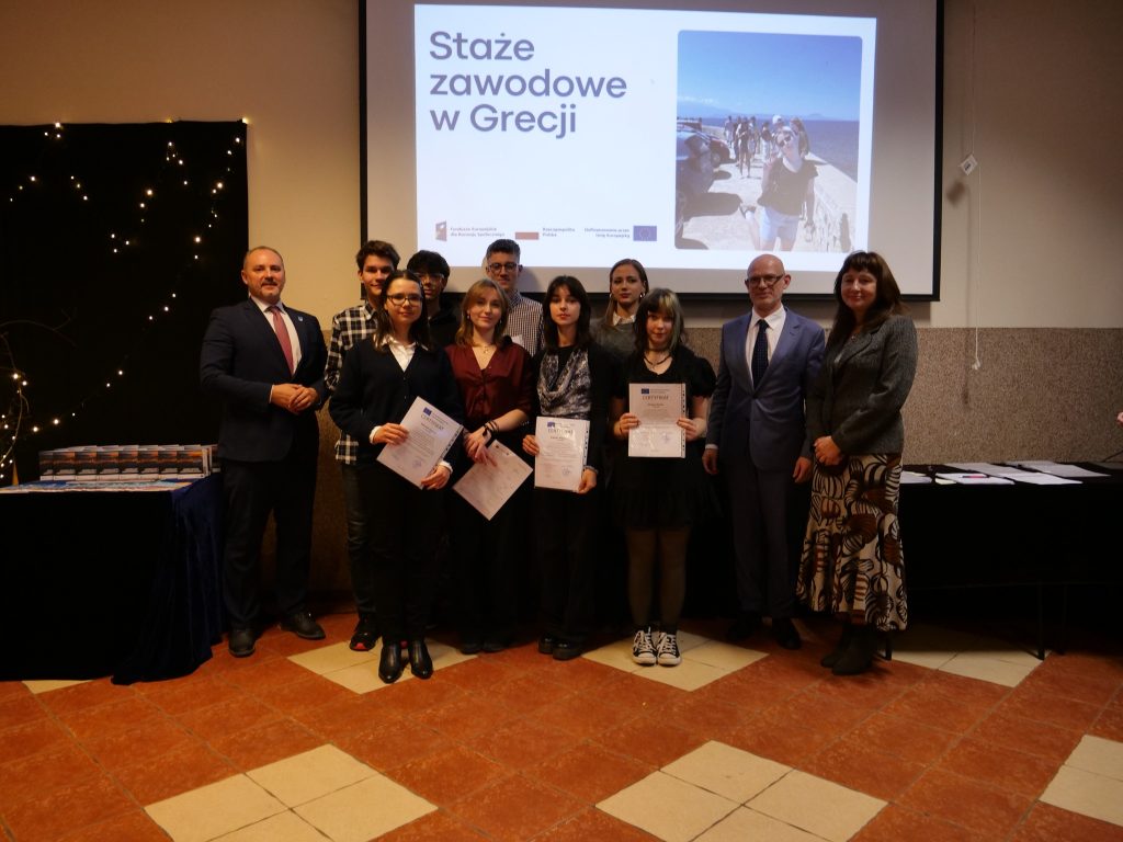 dwóch mężczyzn w garniturze i kobieta pozują do zdjęcia z uczniami którzy w rękach trzymają dyplomy, w tle ekran multimedialny na którym odtwarzana jest kolorowa prezentacja