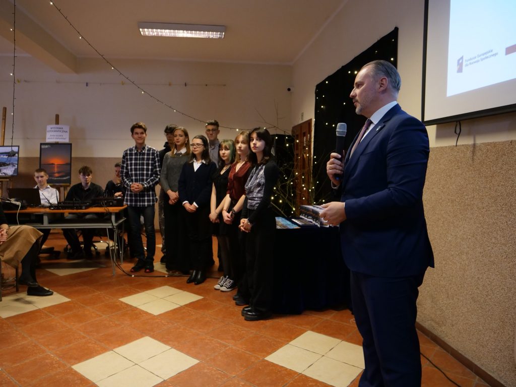 po prawej stronie mężczyzna w garniturze mówi do mikrofonu, w oddali stoi grupa uczniów, obok siedzą uczniowie za konsolą