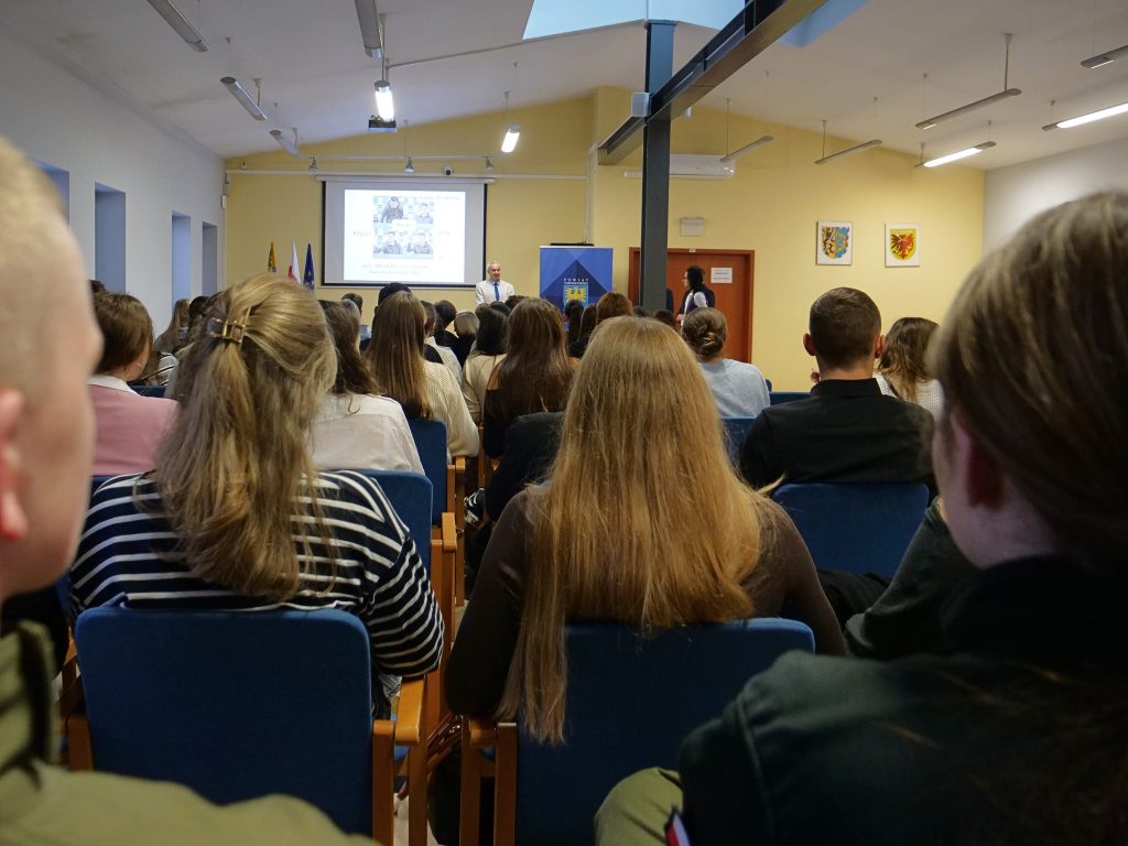 grupa osób siedzi na widowni, na sali sesyjnej, wszyscy są odwróceni tyłem w oddali w tle ekran multimedialny