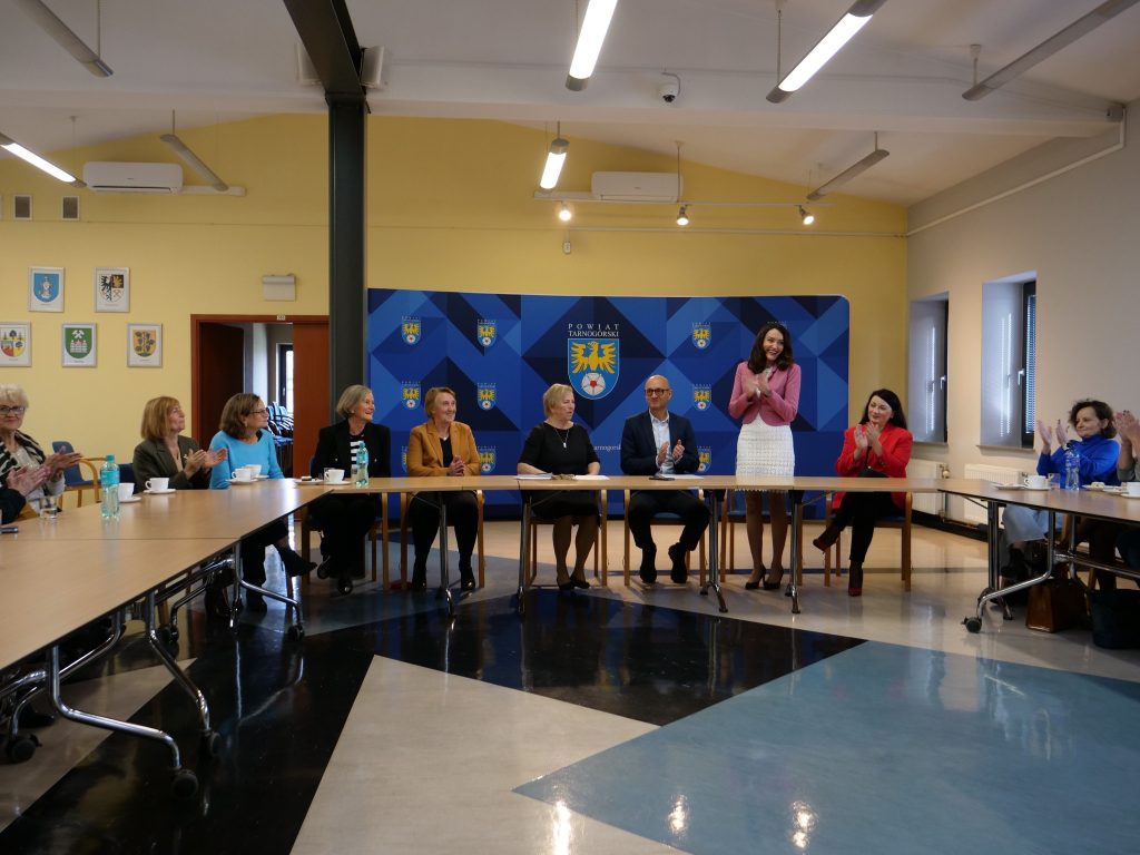 w sali, grupa osób siedzi przy stole, jedna osoba stoi, ma złożone ręce, w tle ścianka z herbem powiatu tarnogórskiego, okna i drzwi