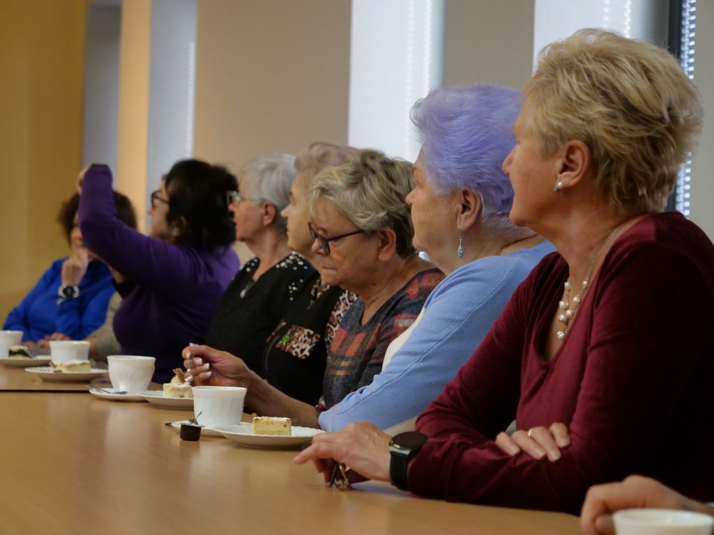 mała grupa kobiet, elegancko ubrane, siedzą przy stole, wszystkie zwrócone w lewą strone