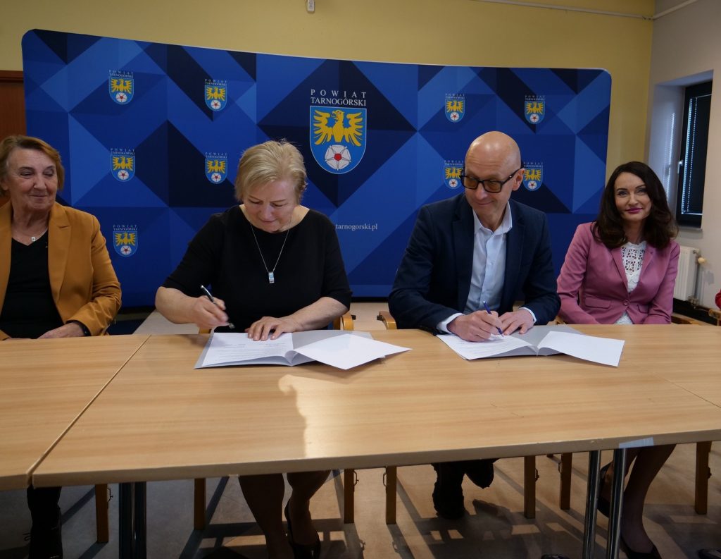 trzy kobiety i mężczyzna siedzą przy stole, kobieta i mężczyzna podpisują dokumenty, w tle ścianka z herbem powiatu tarnogórskiego