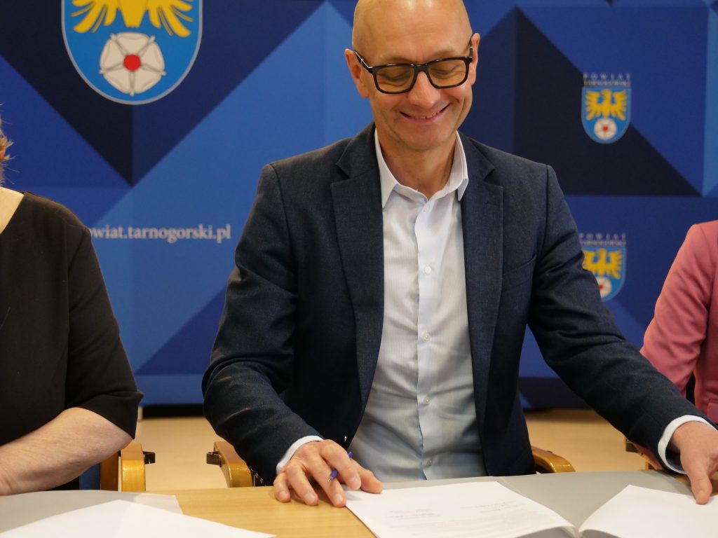 mężczyzna, przy stole siedzi, uśmiecha się do dokumentów w tle ścianka z herbem powiatu tarnogórskiego