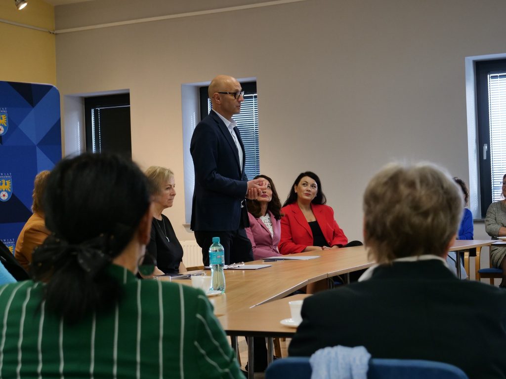 grupa osób siedzi przy stole, jeden mężczyzna stoi, ma złożone ręce, w tle okna z żaluzjami