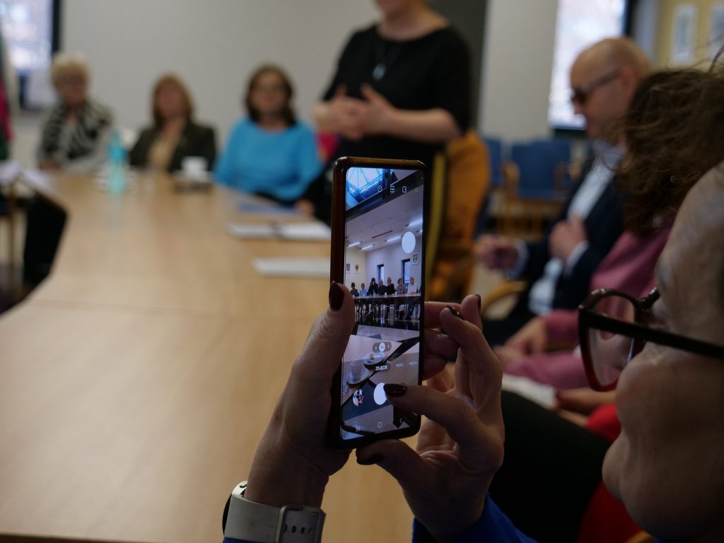 kobieta, robi zdjęcie telefonem, w oddali widać inne osoby, jedna z osób stoi i ma założone ręce 