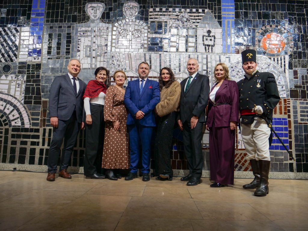 grupa dziewięciu osób, ubrani w stroje okolicznościowe, pozują do zdjęcia, wewnątrz budynku, stoją na tle artystycznej mozaiki, po prawej stronie stoi mężczyzna w stroju górniczym