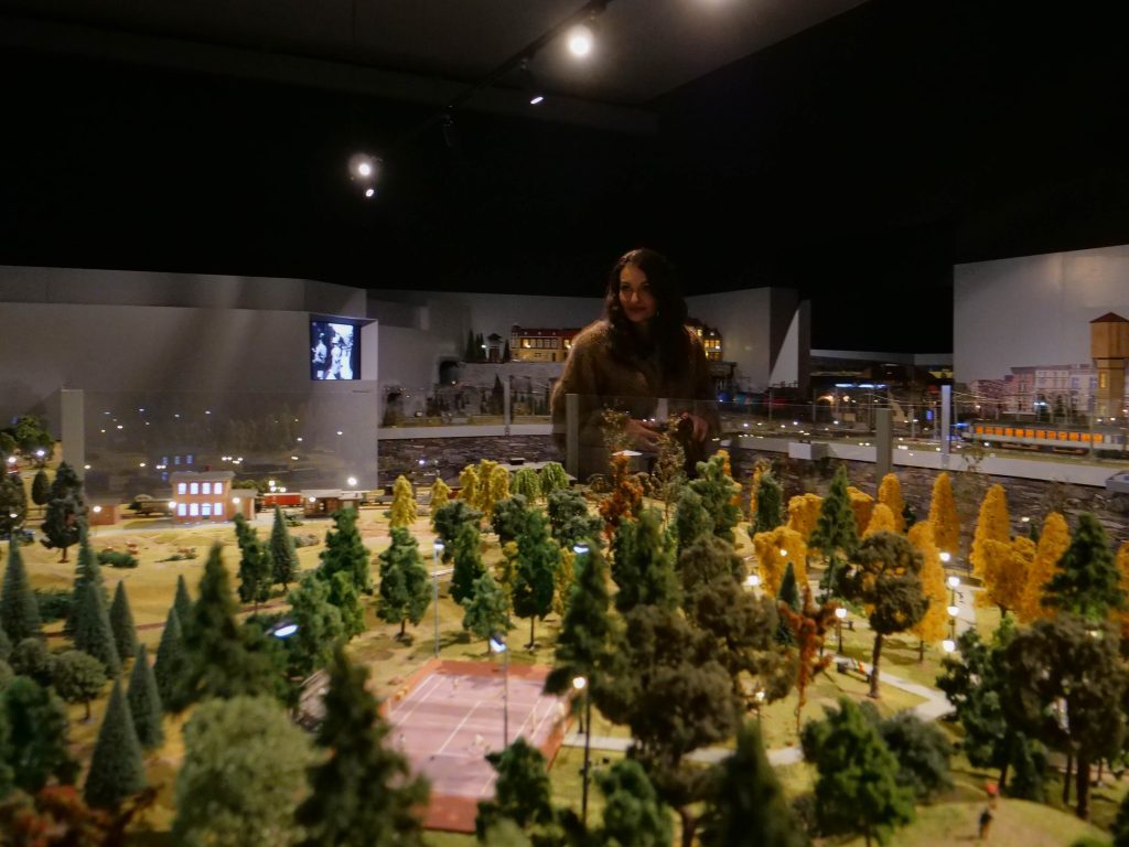 na pierwszym planie w wystawowym muzeum makieta, z dużymi drzewami, w środku boisko do gry, w tle kobieta ubrana w futro, za nią inne makiety