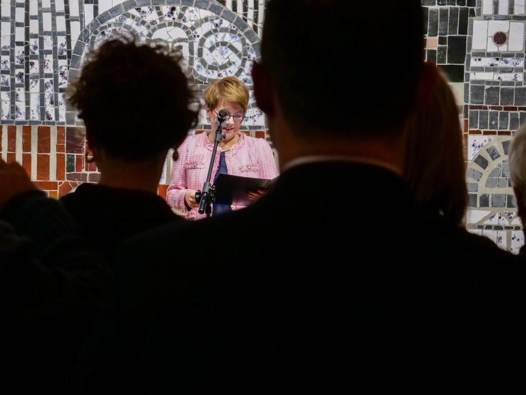 grupa osób, stoi tyłem, spoglądają w przód na kobietę która stoi na scenie,, przed sobą ma mikrofon, ma spuszczoną głowę w dół