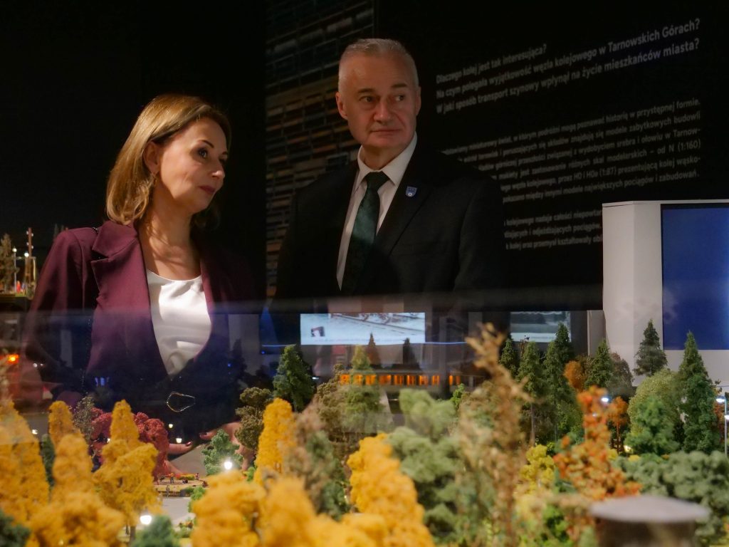 kobieta i mężczyzna, elegancko ubrani, spoglądają przed siebie, na pierwszym planie makieta wystawowa z drzewami