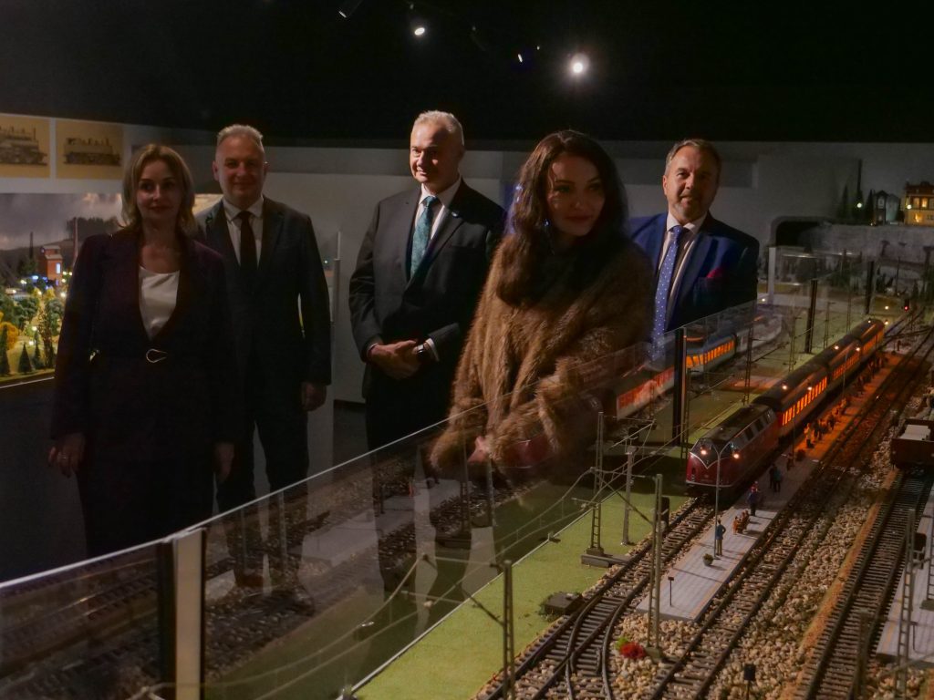 grupa pięciu osób, elegancko ubranych, stoi na sali wystawowej w muzeum, oglądają makietę wystawową, na pierwszym planie makieta torów kolejowych z pociągiem i peronem, w tle elementy makiety miasta i światło ekspozycyjne 