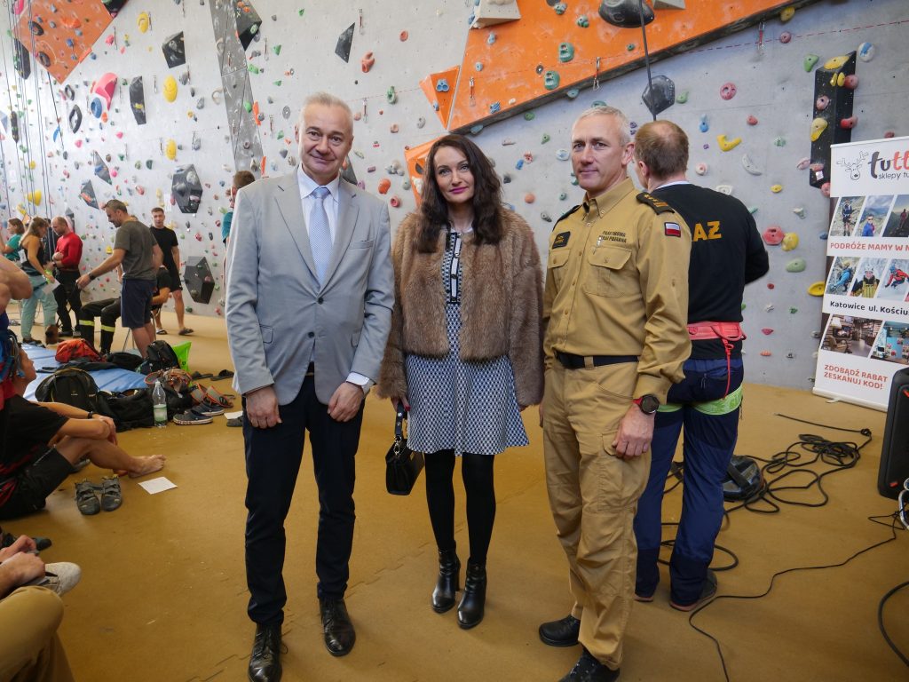 mężczyzna w garniturze, kobieta elegancko ubrana i mężczyzna w mundurze strażackim, stoją na sali ścianki wspinaczkowej, w tle wspinające się osoby