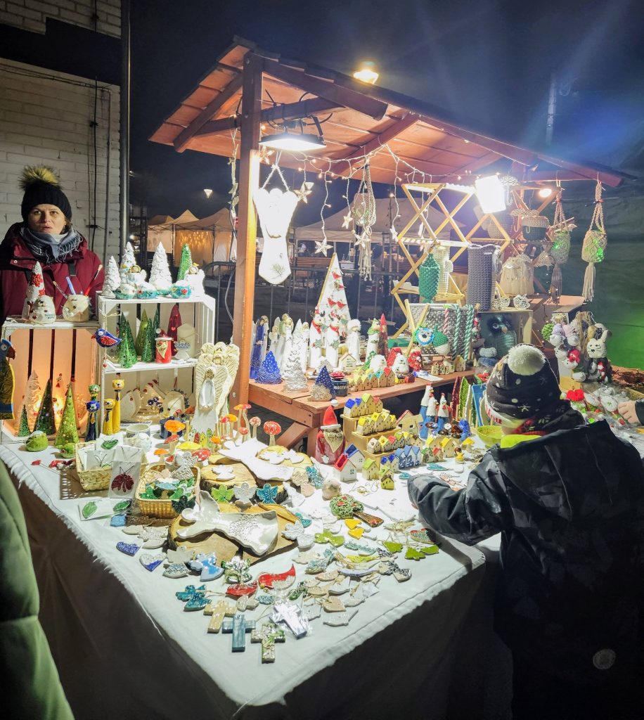 na pierwszym planie bibeloty, ozdoby i dekoracje rozłożone na podwyższeniu, stoisko z drewnianym zadaszeniem - stragan, na drugim planie po lewej kobieta, ubrana w kurtkę, czapkę i szal, po prawej, w dolnym roku, dziecko również w zimowych ubraniach
