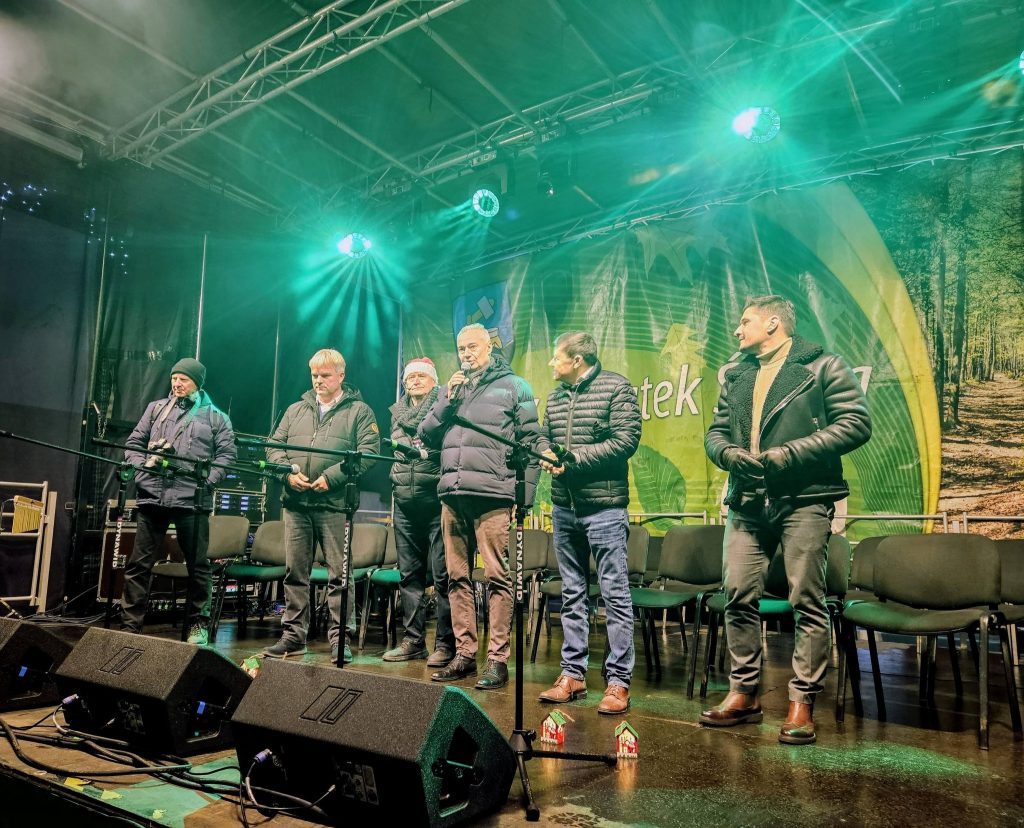 sześciu mężczyzn stoi, przed nimi mikrofony na statywach, za nimi krzesła ustawione rzędami, za krzesłami plakat, powyżej lampy sceniczne i szkielet zadaszenia, przed nimi kolumny odsłuchowe