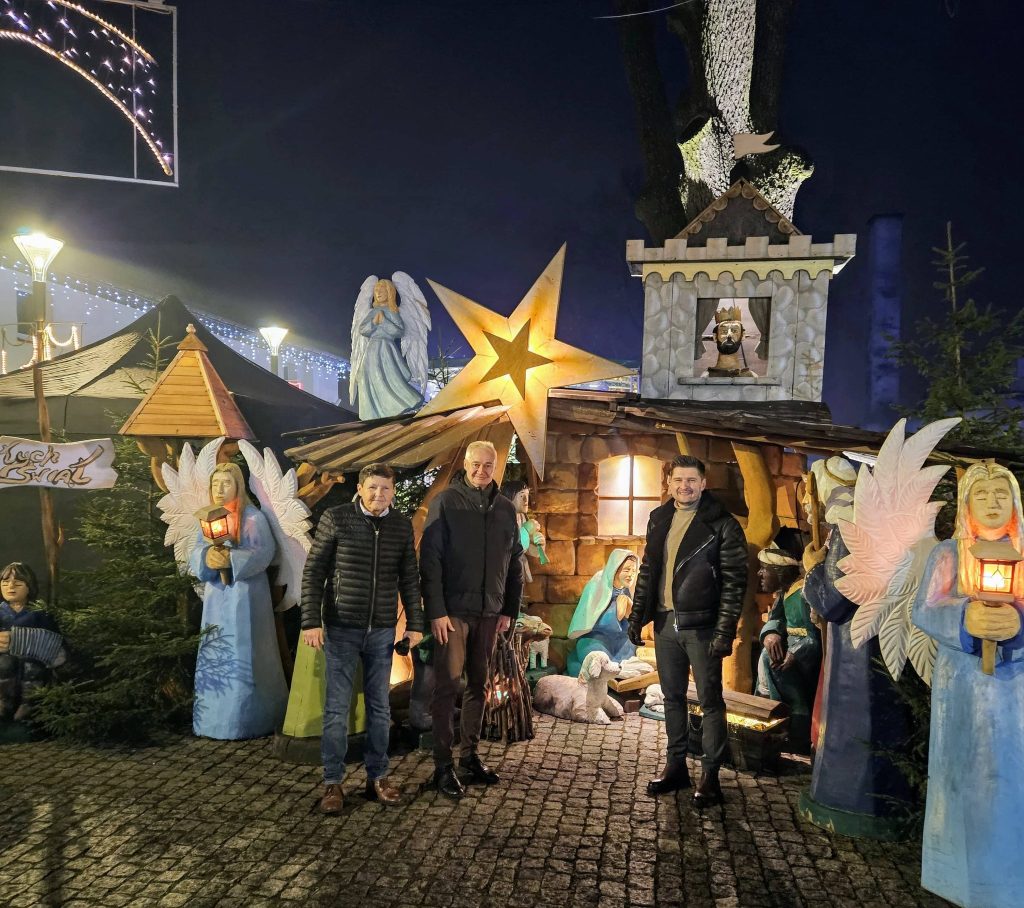 na pierwszym planie trzech mężczyzn, za nimi szopka bożonarodzeniowa, figury aniołów, gwiazda betlejemska, powyżej nocne niebo