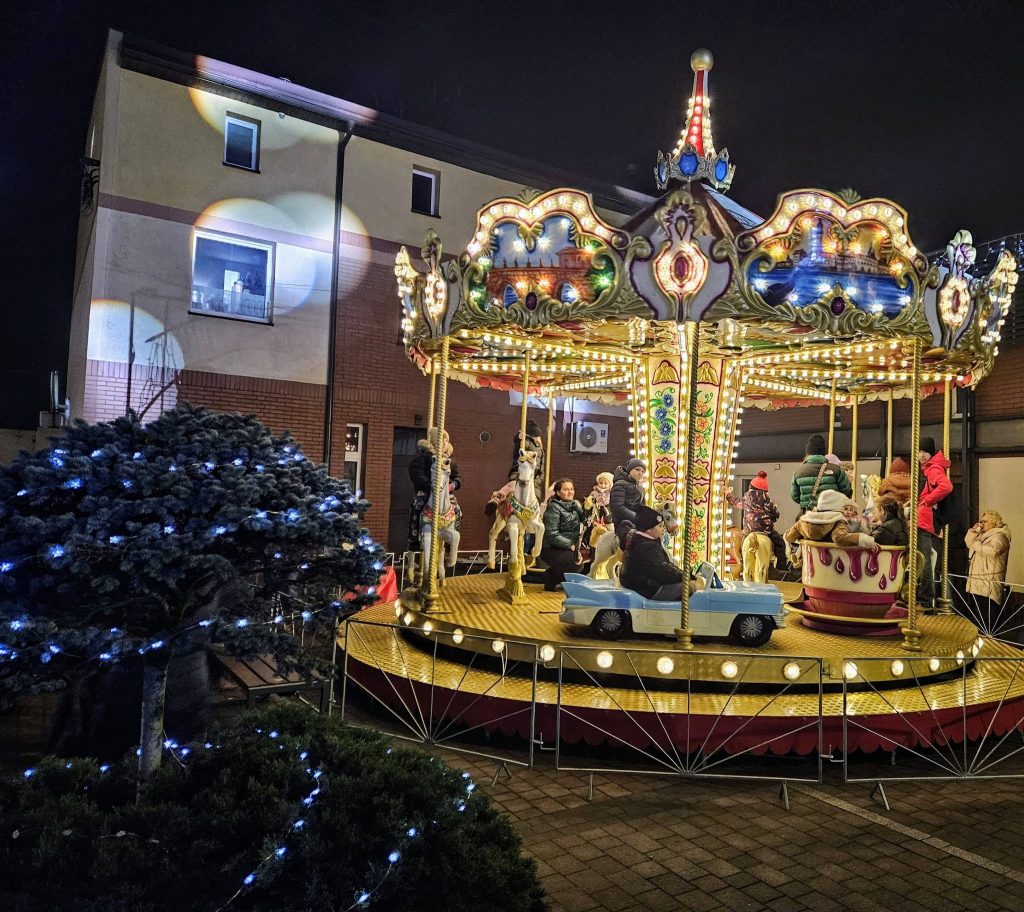po lewej fragment rośliny oplecionej lampkami dekoracyjnymi, pośrodku karuzela obrotowa, za nią fragment budynku