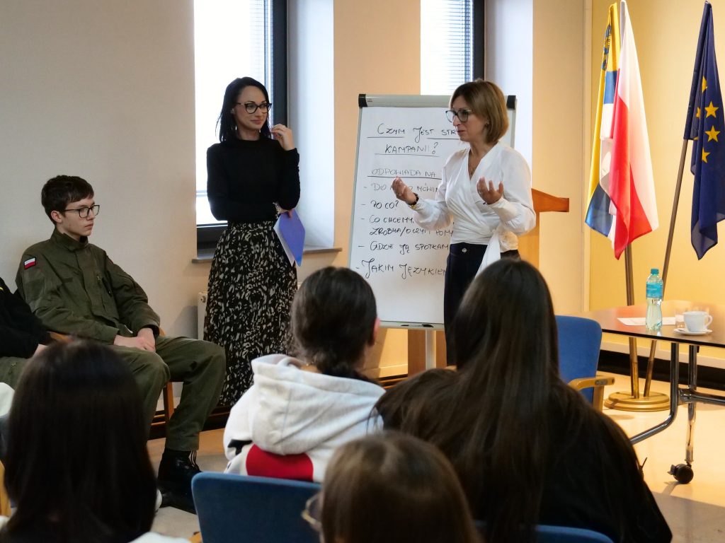 na pierwszym planie dwie kobiety prowadzące, stoją przed grupą młodzieży i omawiają zagadnienia, gestykulują, między nimi tablica z zapisanymi słowami, obok stolik z materiałami, w tle flagi, uczestnicy siedzą na krzesłach