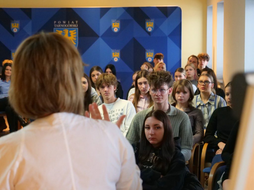 na pierwszym planie odwrócona tyłem, przodem do młodzieży, młodzież siedzi na krzesłach, wsłuchana w wykład , w tle ścianka z herbem powiatu